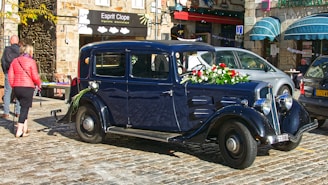 A vintage black car is adorned with flowers, including red roses and green foliage, parked on cobblestone in a charming street setting. Nearby, people walk past and cafes with awnings add a lively atmosphere.