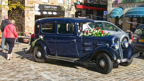 A vintage black car is adorned with flowers, including red roses and green foliage, parked on cobblestone in a charming street setting. Nearby, people walk past and cafes with awnings add a lively atmosphere.