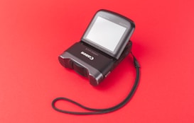 A digital camera lies on a vivid red background. The camera has a flip-up screen and a visible wrist strap, adding a practical feature for secure handling. The lens is facing down, emphasizing the compact design of the device.