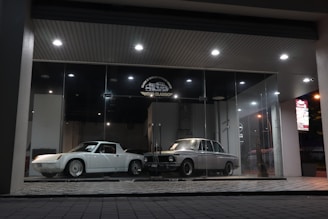 A pair of classic cars are displayed in an illuminated glass showroom. One car is white and sleek with angular features, while the other is silver-gray with a vintage boxy design. The floor is a checkered tile pattern, and spotlights from the ceiling cast a soft glow on the cars.