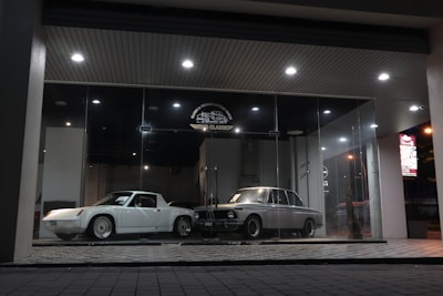 A pair of classic cars are displayed in an illuminated glass showroom. One car is white and sleek with angular features, while the other is silver-gray with a vintage boxy design. The floor is a checkered tile pattern, and spotlights from the ceiling cast a soft glow on the cars.