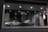 A pair of classic cars are displayed in an illuminated glass showroom. One car is white and sleek with angular features, while the other is silver-gray with a vintage boxy design. The floor is a checkered tile pattern, and spotlights from the ceiling cast a soft glow on the cars.