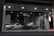 A pair of classic cars are displayed in an illuminated glass showroom. One car is white and sleek with angular features, while the other is silver-gray with a vintage boxy design. The floor is a checkered tile pattern, and spotlights from the ceiling cast a soft glow on the cars.