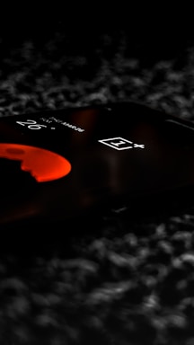 A smartphone screen displays a digital clock with a partially visible number and logo, illuminated in a dim setting. The design incorporates a sleek, modern appearance with an emphasis on minimalism. The background is blurred, enhancing the focus on the phone.
