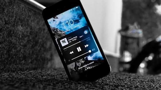 A smartphone displaying a music player interface rests on a textured surface. The screen shows a blue sky and clouds background with music controls and the time and date. The device is tilted and positioned near a staircase.