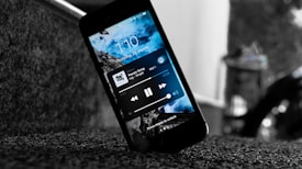 A smartphone displaying a music player interface rests on a textured surface. The screen shows a blue sky and clouds background with music controls and the time and date. The device is tilted and positioned near a staircase.