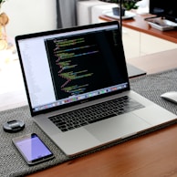 A modern laptop displaying code is placed on a wooden desk, accompanied by a smartphone, a lens cap, and a computer mouse. The background shows a part of a living room with a piece of furniture and a blurred out television. The workspace setup is neat and organized, suggesting a focus on productivity.