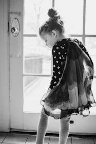 grayscale photo of girl in polka dot dress looking out the window