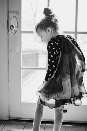 A young girl stands near a glass door, looking down pensively with her hair in a bun. She is wearing a dress with butterfly wing designs, creating a whimsical effect. The setting appears calm and the lighting is soft, highlighting her contemplative mood.