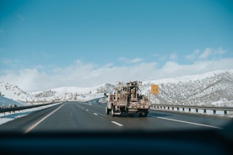 A highway scene featuring a large truck carrying equipment traveling on a multi-lane road through a snow-covered mountainous area. The sky is clear with a few clouds, and there is a road sign indicating an exit with a speed limit of 35 mph.