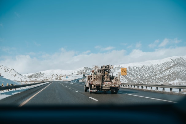 A highway scene featuring a large truck carrying equipment traveling on a multi-lane road through a snow-covered mountainous area. The sky is clear with a few clouds, and there is a road sign indicating an exit with a speed limit of 35 mph.