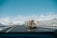 A highway scene featuring a large truck carrying equipment traveling on a multi-lane road through a snow-covered mountainous area. The sky is clear with a few clouds, and there is a road sign indicating an exit with a speed limit of 35 mph.