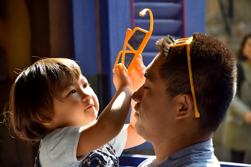 A child playfully interacts with an adult, holding up a pair of orange plastic glasses. The adult has a similar pair placed on their head, and both appear to be sharing a warm, affectionate moment. Soft, natural light bathes their faces, highlighting their expressions.