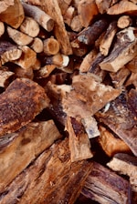 A pile of chopped firewood, featuring various sizes and shapes of logs. The wood displays rough, textured surfaces with visible bark and grain patterns. The logs are primarily brown with some darker and lighter shades.