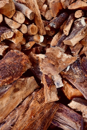 A pile of chopped firewood, featuring various sizes and shapes of logs. The wood displays rough, textured surfaces with visible bark and grain patterns. The logs are primarily brown with some darker and lighter shades.
