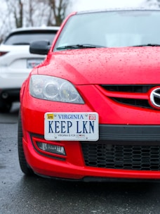 A red car with a Virginia license plate that reads 'KEEP LKN' is parked on wet pavement. The car is positioned in front of a white vehicle, and raindrops are visible on the surface of the red car.