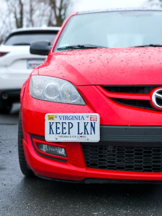 A red car with a Virginia license plate that reads 'KEEP LKN' is parked on wet pavement. The car is positioned in front of a white vehicle, and raindrops are visible on the surface of the red car.
