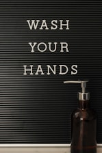 A black letter board with white letters spelling the phrase 'WASH YOUR HANDS' is mounted on the left side. On the right, there is a dark brown soap dispenser with a silver pump resting on a white surface.