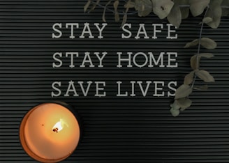 A black letter board displays the message 'STAY SAFE STAY HOME SAVE LIVES.' A lit candle is positioned in the lower left corner, casting a warm glow. Dried eucalyptus leaves hang from the top right corner, adding a natural element.