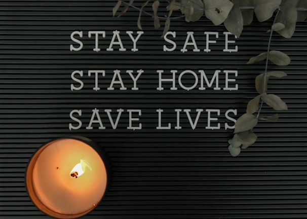 A black letter board displays the message 'STAY SAFE STAY HOME SAVE LIVES.' A lit candle is positioned in the lower left corner, casting a warm glow. Dried eucalyptus leaves hang from the top right corner, adding a natural element.