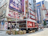 A Coca-Cola delivery truck is parked on a busy street next to a building covered with colorful anime posters. Several cardboard boxes are stacked around the truck, and a man is seen near the truck, possibly handling deliveries. Pedestrians are walking along the street surrounded by tall buildings with vibrant signage.