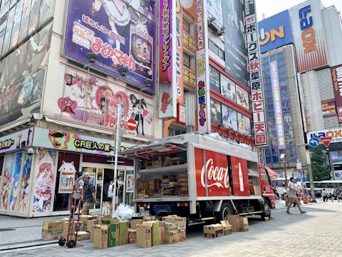 A Coca-Cola delivery truck is parked on a busy street next to a building covered with colorful anime posters. Several cardboard boxes are stacked around the truck, and a man is seen near the truck, possibly handling deliveries. Pedestrians are walking along the street surrounded by tall buildings with vibrant signage.