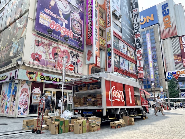 A Coca-Cola delivery truck is parked on a busy street next to a building covered with colorful anime posters. Several cardboard boxes are stacked around the truck, and a man is seen near the truck, possibly handling deliveries. Pedestrians are walking along the street surrounded by tall buildings with vibrant signage.