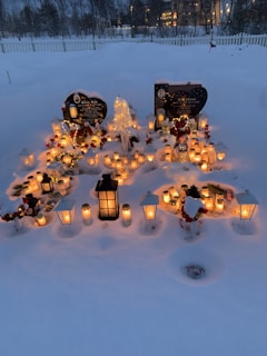 A snow-covered grave site adorned with numerous candles and lanterns, creating a warm glow against the snowy background. There are two headstones visible, surrounded by floral arrangements and winter decorations.