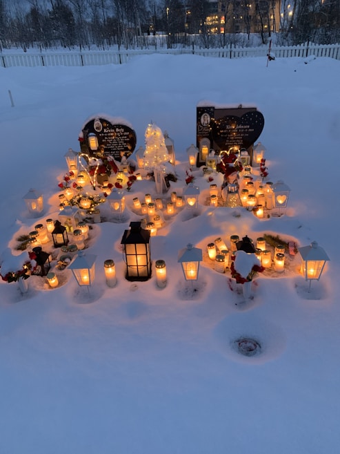 A snow-covered grave site adorned with numerous candles and lanterns, creating a warm glow against the snowy background. There are two headstones visible, surrounded by floral arrangements and winter decorations.
