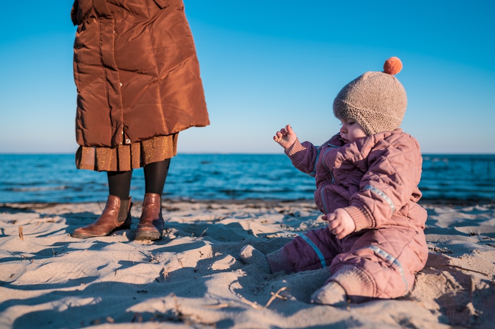 child in brown jacket and gray knit cap sitting on white snow covered ground during daytime