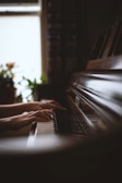 Hands are playing on a piano keyboard. The lighting is soft, creating a warm, calm atmosphere, with plants visible in the blurred background near a window.