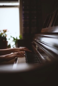 Hands are playing on a piano keyboard. The lighting is soft, creating a warm, calm atmosphere, with plants visible in the blurred background near a window.