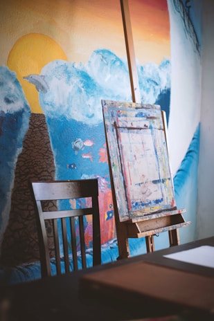 An artist's workspace is depicted, featuring an easel with a blank canvas and a wooden chair nearby. The background showcases a mural of ocean waves under a blue sky, with the yellow sun setting in the distance, casting a warm glow. The mural is vibrant with various shades of blue, and small painted fish add detail.