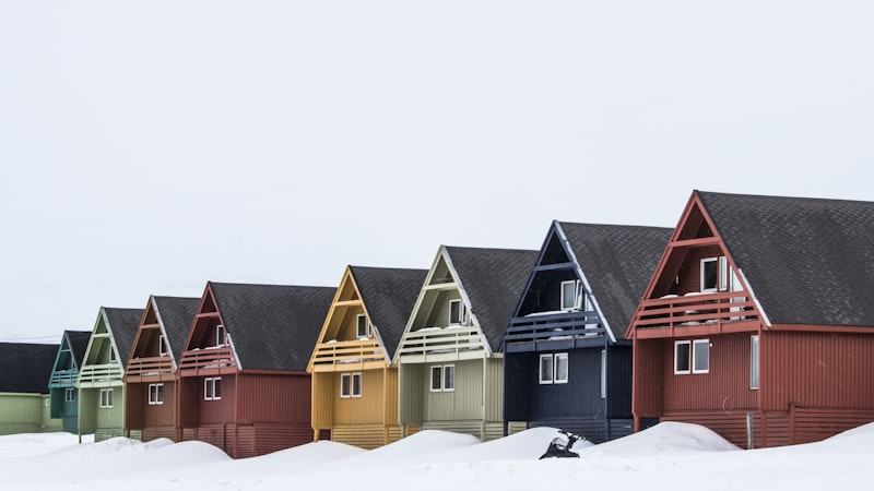 Row of A-frame wooden houses painted red, blue, yellow, green, and teal standing in deep snow under a flat white sky