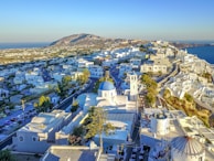 A scenic view of a coastal Greek town with white-washed buildings spread across a hillside. The landscape includes winding streets, a church with a blue dome and bell tower, and the blue sea in the background under a clear sky. There is a mountain in the distance on the left side.