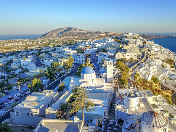 A scenic view of a coastal Greek town with white-washed buildings spread across a hillside. The landscape includes winding streets, a church with a blue dome and bell tower, and the blue sea in the background under a clear sky. There is a mountain in the distance on the left side.