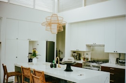 A modern kitchen with white cabinetry and a large central island topped with marble. Four woven wooden chairs are positioned along the island. A stylish, wide-mesh pendant light hangs above. On the counter, there is a vase with flowers, bottles, and kitchen essentials. The back wall features a stainless steel stove with a matching backsplash, and a sleek stainless refrigerator is tucked into a cabinet alcove.