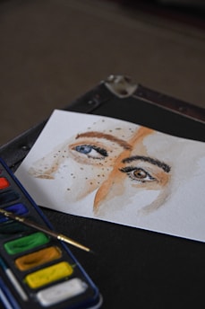 A watercolor painting on a white sheet portrays the close-up of two eyes, one blue and one brown, with freckles scattered across the bridge of the nose. The painting is placed on a dark surface next to a palette containing several colors and a paintbrush.