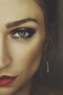 A close-up of the left side of a woman's face highlighting a striking blue eye with bold eye makeup, including dark eyeshadow and mascara. Her eyebrow is well-defined, and she wears bright red lipstick. The image captures a sense of intense focus and dramatic style.