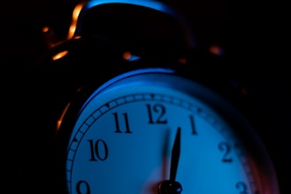 A close-up view of an analog alarm clock with a dark background and ambient blue lighting. The clock face is partially illuminated, showing the numbers clearly.