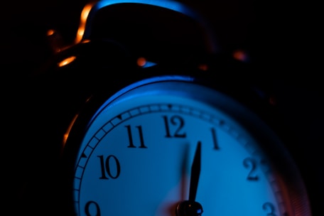 A close-up view of an analog alarm clock with a dark background and ambient blue lighting. The clock face is partially illuminated, showing the numbers clearly.