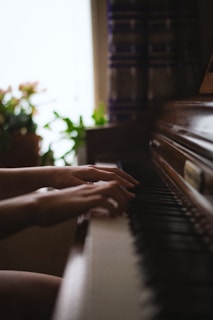 Hands gracefully playing the keys of a piano, with soft lighting casting gentle shadows. A blurred background reveals a window with light streaming in, highlighting some potted plants. The setting appears calm and intimate.