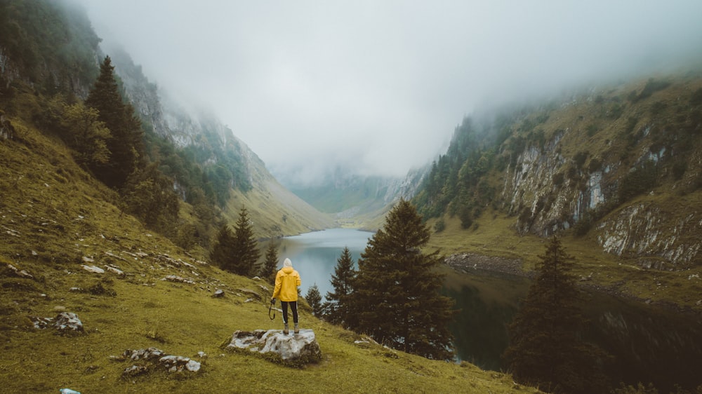 person in yellow jacket and black pants standing on rock near river during daytime