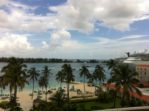 A tropical beach scene features tall palm trees along the shoreline and a serene, sandy beach. Lounge chairs with red umbrellas are scattered across the sand. Beyond the beach, a large cruise ship is docked in the calm blue ocean, with a backdrop of lush greenery on a distant island. The sky is partly cloudy, adding a relaxed and inviting atmosphere.