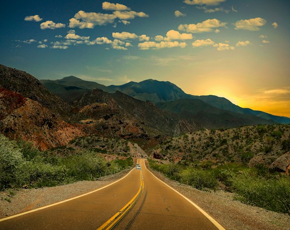 A long, winding road stretches into a mountain range under a sky dotted with fluffy clouds. The sun casts a warm glow over the landscape, illuminating the rugged terrain and sparse vegetation along the roadside. A solitary vehicle travels down the highway, adding a sense of scale to the vast and expansive scene.
