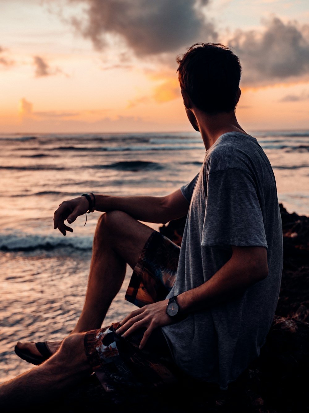 Hombre en camiseta gris sentado en roca marrón cerca del mar durante la puesta del sol