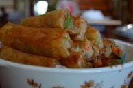 A pile of freshly fried spring rolls is placed in a decorative bowl. The rolls have a golden-brown crispy texture and some visible vegetable fillings, such as carrots and green herbs. The image conveys a sense of homemade cooking and comfort food.