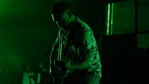 A silhouetted musician playing an electric guitar is bathed in green light. The background is dark, with a few indistinct shapes, possibly other band members or equipment.
