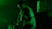 A silhouetted musician playing an electric guitar is bathed in green light. The background is dark, with a few indistinct shapes, possibly other band members or equipment.