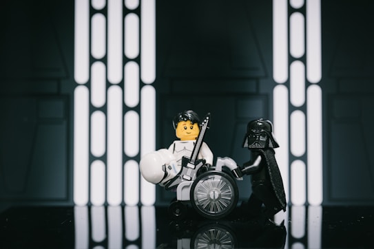 Two LEGO figures engage in a playful scene set against a futuristic backdrop. One figure in a black outfit appears to assist or confront another figure with shaggy hair, holding a guitar-like object. The set design evokes a sci-fi atmosphere with geometric patterns and glowing elements.